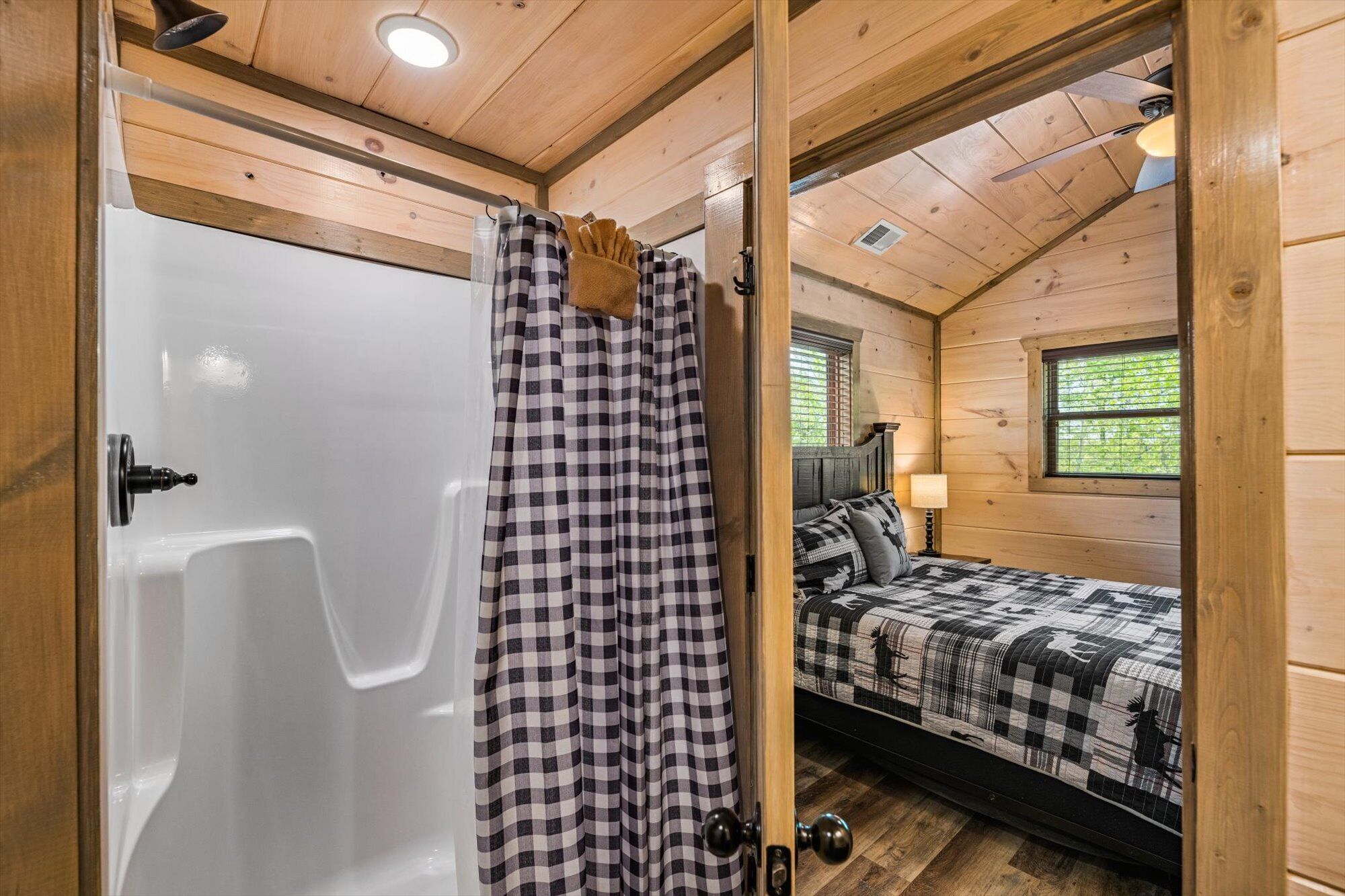 A wooden cabin room with a white bathtub, checkered shower curtain, and a bed with a checkered bedspread.