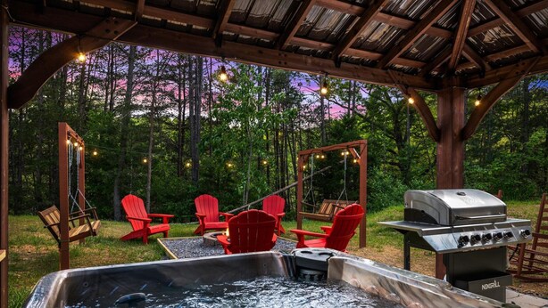 A hot tub with a grill, red Adirondack chairs, and a swing set in a wooded area. - NEW Cabin 10mins to Dollywood★HotTub★BBQ★GSMNP★PetsOK★Highspeed WiFi★PvtBackyard (Pigeon Forge)