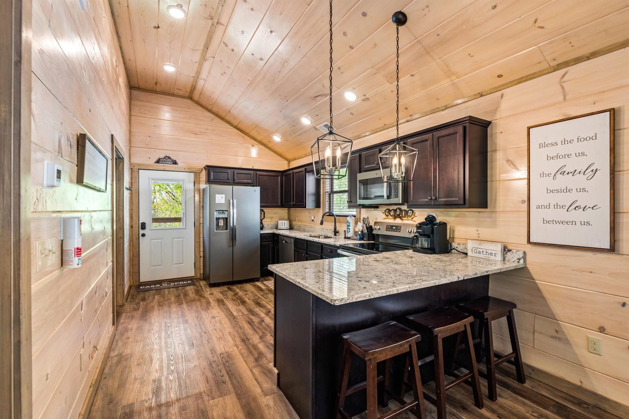 A kitchen with wooden walls, a central island with stools, dark cabinets, stainless steel appliances, and a ceiling with recessed lighting.