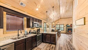 A cabin-style kitchen with wooden walls, dark cabinets, granite countertops, and a stone fireplace. - NEW Cabin 10mins to Dollywood★HotTub★BBQ★GSMNP★PetsOK★Highspeed WiFi★PvtBackyard (Pigeon Forge)