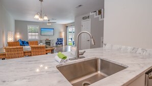A modern kitchen with a large sink, a dining area with wicker furniture, and a view of the outdoors through large windows.