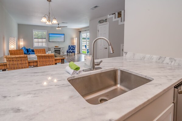 A modern kitchen with a large sink, a dining area with wicker furniture, and a view of the outdoors through large windows.