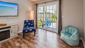 A living room with a fireplace, a television, a star-patterned chair, a bean bag, and a sliding glass door leading to a patio with blue chairs.
