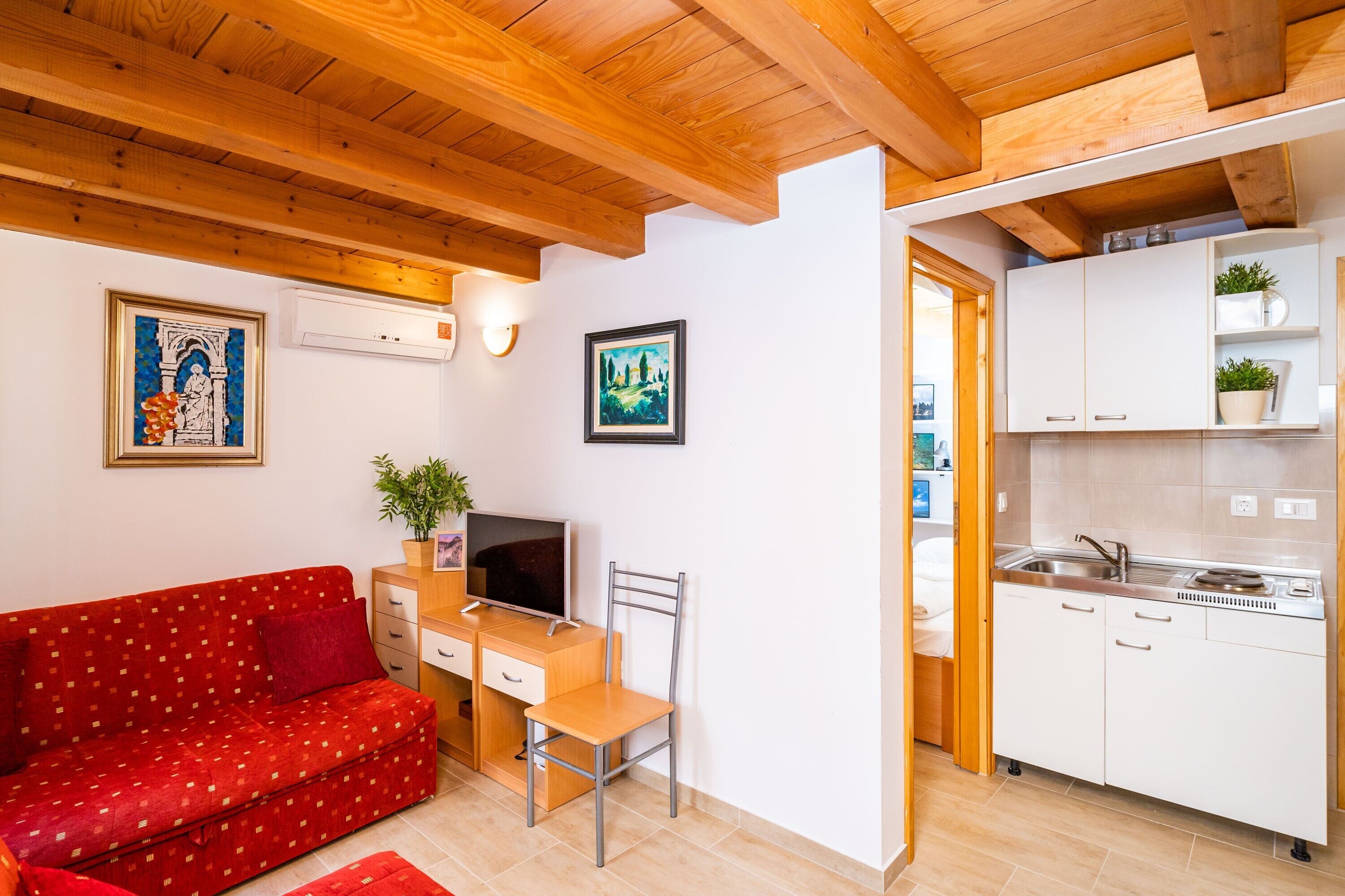 A compact living space with a red sofa, a wooden desk with a chair, a television, and a kitchenette with white cabinets and a sink.