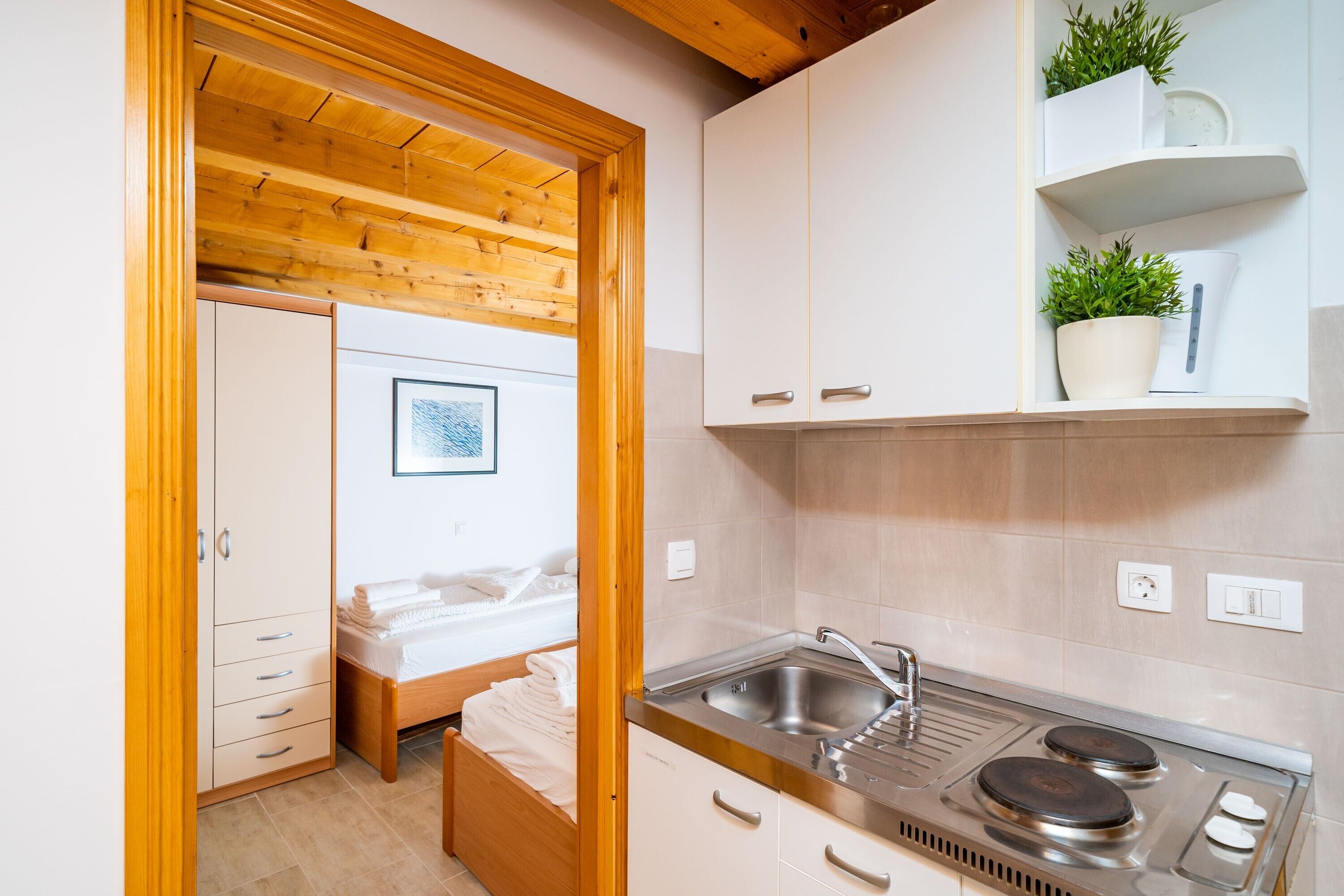 Une cuisine compacte équipée d’un évier, d’une cuisinière et de placards. On y trouve un lit avec une tête de lit blanche et une petite table de chevet. Une porte en bois donne accès à une autre pièce.