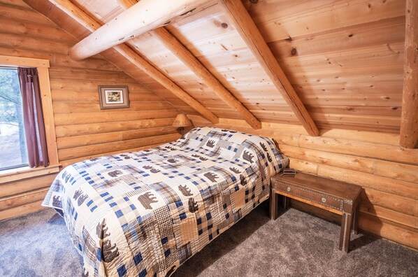 Une chambre confortable dans un chalet, avec un plafond en bois, un lit simple recouvert d’une courtepointe à motifs, une petite table de chevet et un tableau encadré accroché au mur.