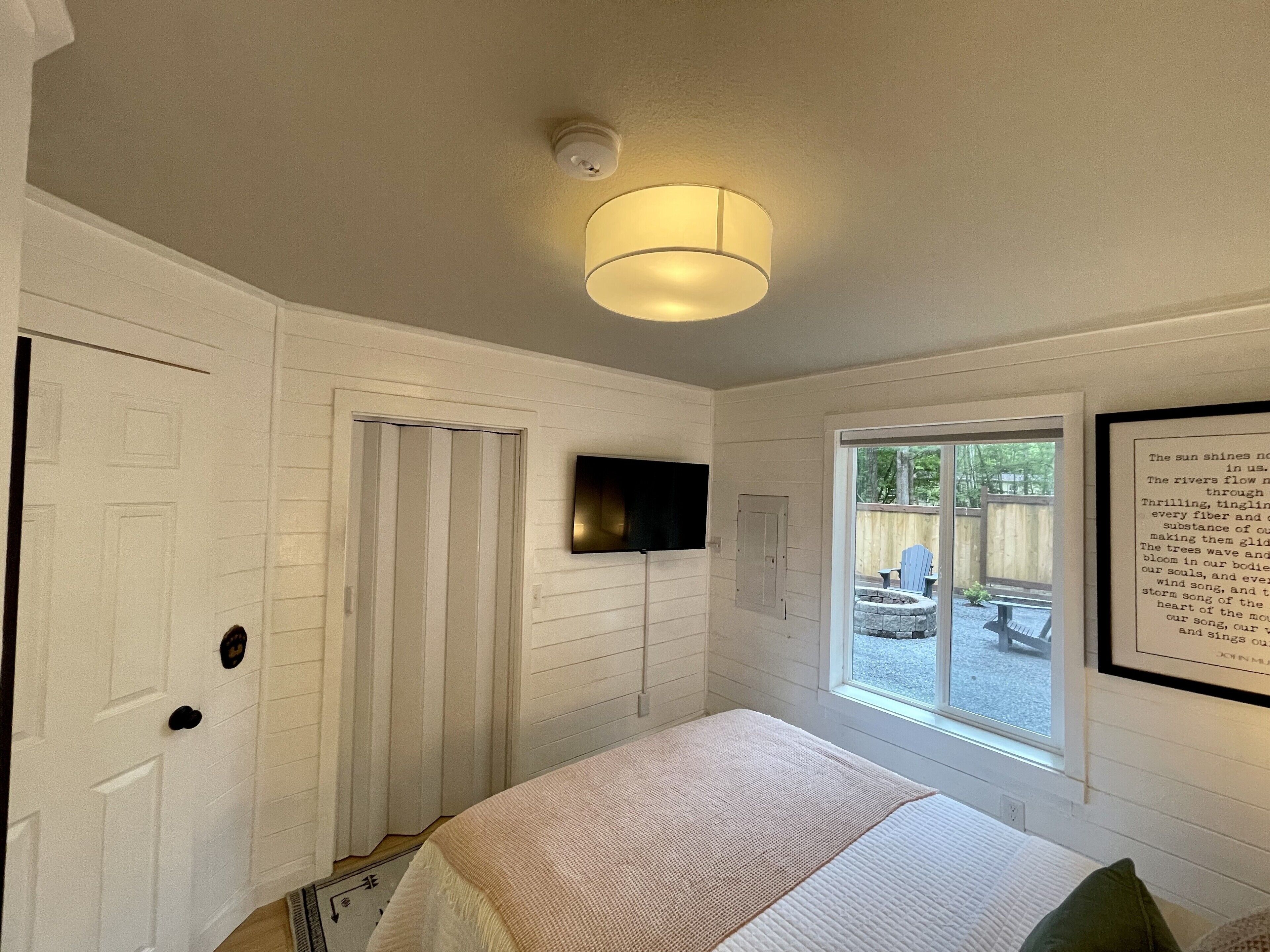 Un dormitorio con cama, televisor montado, ventana con vista al exterior y lámpara en el techo.