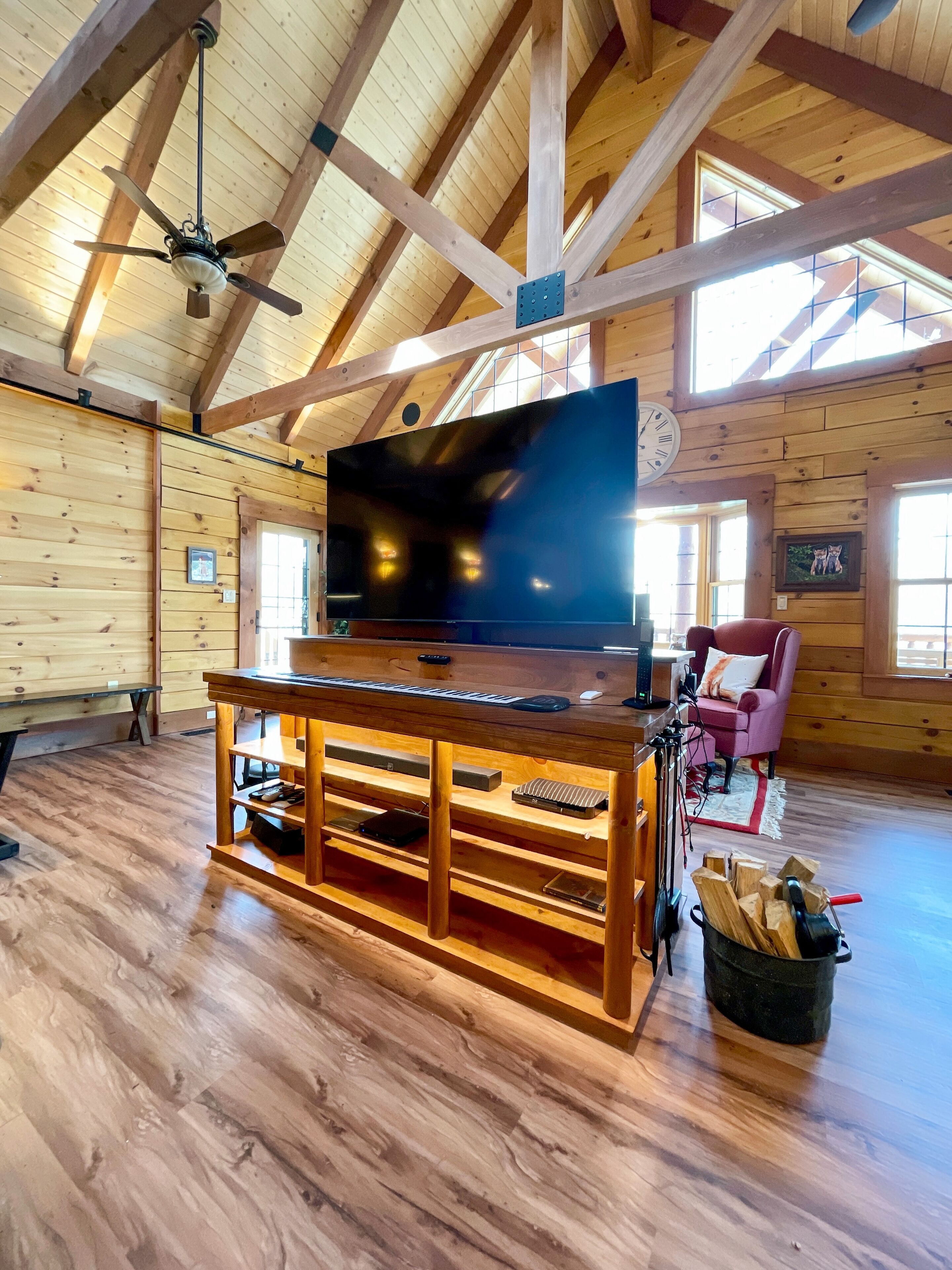 A spacious living room with a large flat-screen TV on a wooden stand, a ceiling fan, and a cozy seating area with a pink chair.