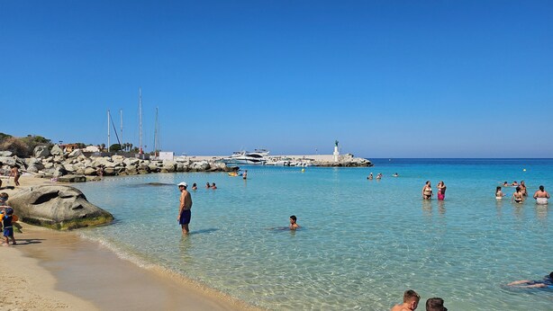 A beach with clear water, people swimming and sunbathing, and yachts docked near a rocky breakwater.