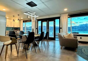 A modern living room with a large window, a dining area with chairs and a table, and a kitchen in the background.