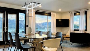 Un espace repas moderne avec une table en verre, des chaises noires et blanches, un luminaire suspendu et un téléviseur fixé au mur.