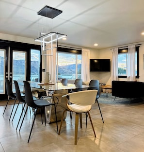 A modern dining area with a glass table, black and white chairs, a hanging light fixture, and a TV mounted on the wall.
