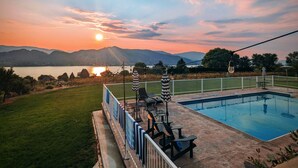 Une piscine entourée de chaises longues et de parasols, elle-même délimitée par une clôture, offrant une vue sur les montagnes et un lac au coucher du soleil.