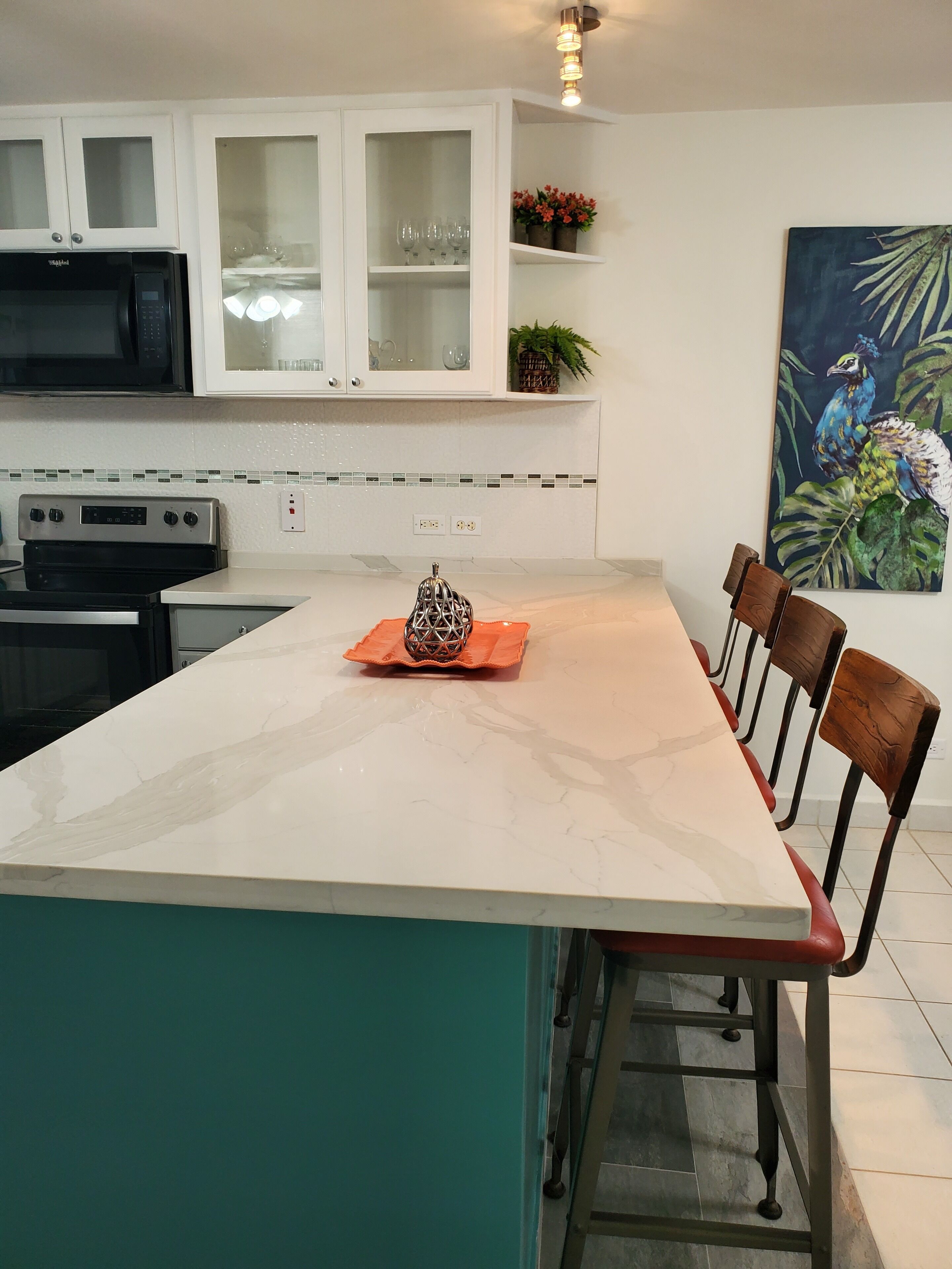 Una cocina con armarios blancos, microondas, horno y comedor con una isla de mármol y sillas de madera.