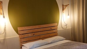 Ein Schlafzimmer mit einem Holz-Kopfteil, einer gemusterten Tagesdecke, einem kleinen Holzschemel und einer an der Wand befestigten Leuchte.