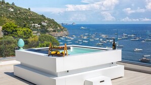 Piscina infinita moderna con terraza de madera y frutero, con vistas a un puerto deportivo con barcos y un paisaje costero.