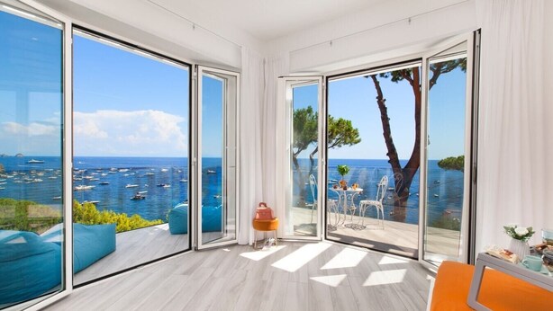 Una habitación moderna con grandes puertas correderas de vidrio que dan a un balcón con vistas al mar y barcos.