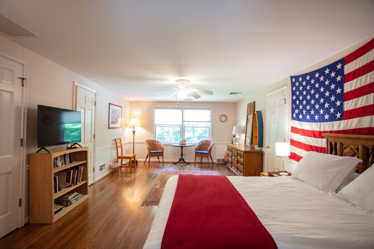 Un dormitorio con una gran bandera estadounidense, una cama, un televisor, una estantería, un escritorio y una ventana con luz natural.
