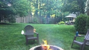 A backyard with a stone fire pit, two Adirondack chairs, and a wooden fence in the background.