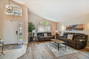 A spacious living room with a brown leather sofa, a coffee table, and a large window with blinds. - Family Friendly Villa Private Pool Game Room Gated Resort Minutes to Disney! (Kissimmee)