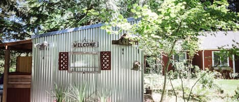 A corrugated metal shed with a 'WELCOME' sign, surrounded by potted plants and trees.