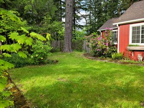 A residential backyard with a red house, a large tree, and various plants and flowers.