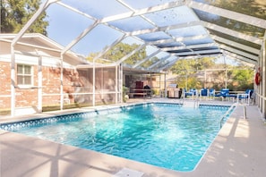 Une piscine couverte avec un toit en verre, entourée d’un mur en briques et d’un espace patio aménagé avec des chaises.