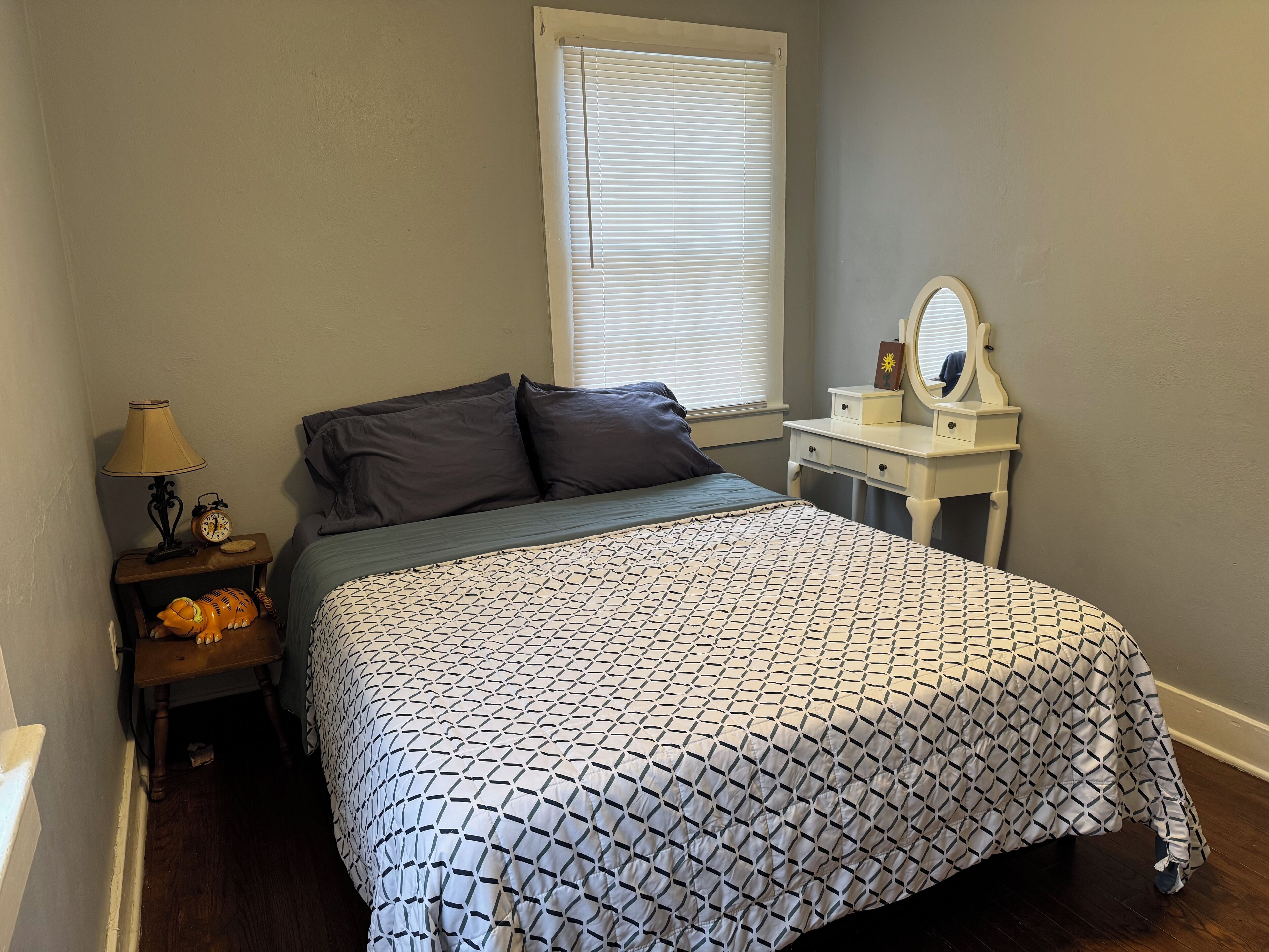 Un dormitorio con cama, mesita de noche, cómoda, espejo y ventana con persianas.