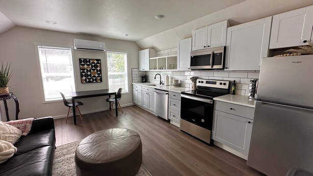 A modern kitchen with white cabinetry, a black countertop, and stainless steel appliances. There is a dining area with a black leather couch and a round ottoman. - Spacious 1bd/1bt downtown apartment next to the park (Vancouver)