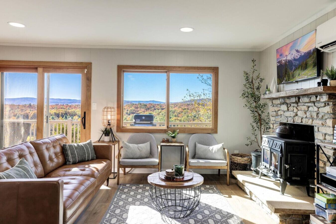 A cozy living room with a leather sofa, two armchairs, a coffee table, and a fireplace. Large windows offer a scenic view of mountains.