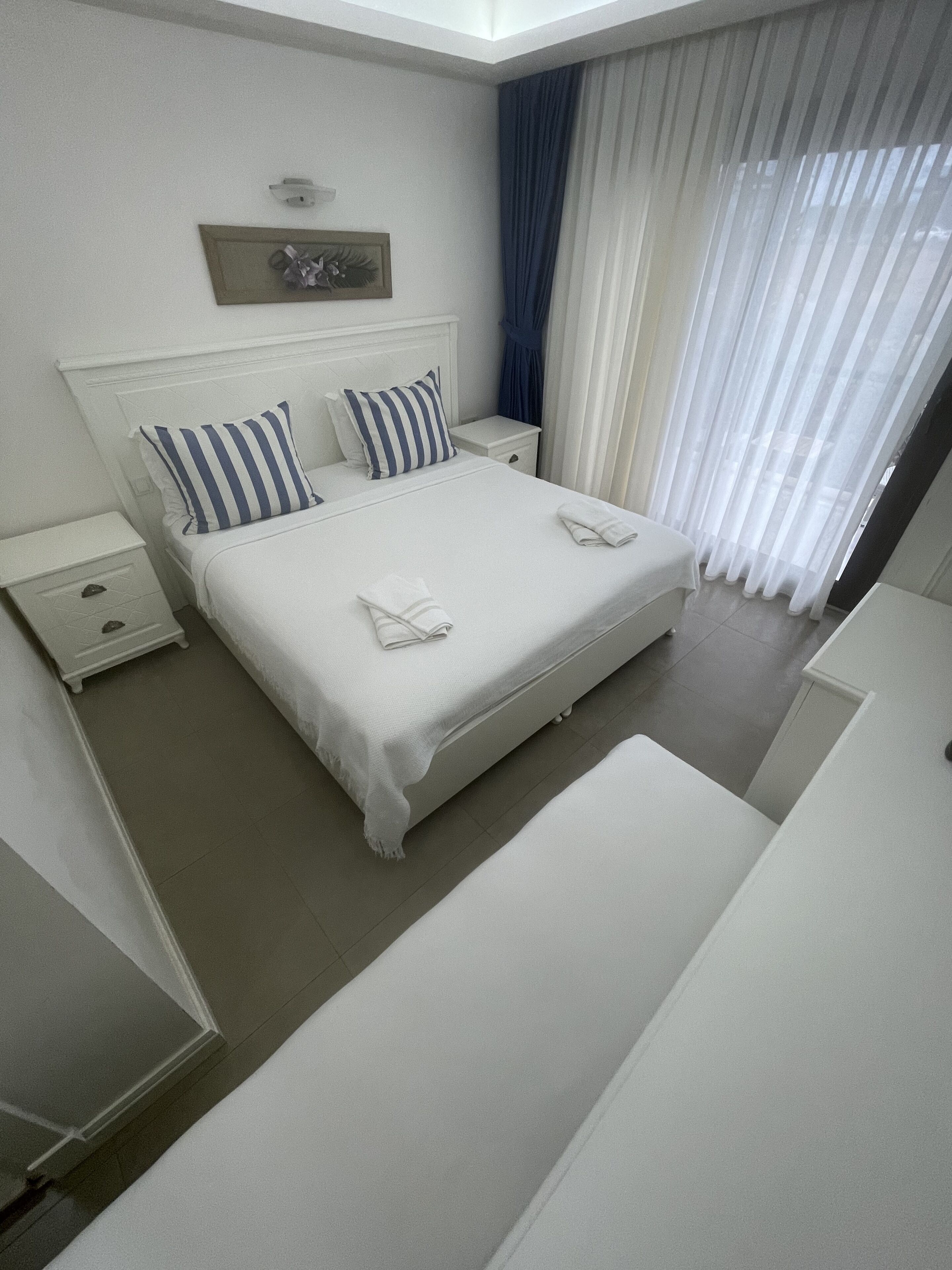 A neatly arranged hotel room with a large bed, white bedding, a nightstand, and a window with sheer curtains.