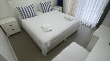 A neatly arranged hotel room with a large bed, white bedding, a nightstand, and a window with sheer curtains.
