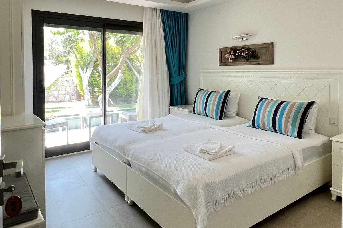 Una cama grande con ropa blanca, una mesita de noche, una lámpara de pared y vistas al exterior.