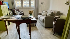 Een moderne woonkamer met een houten eettafel gedekt voor twee personen, een beige bank en een uitzicht op gebouwen door grote ramen.
