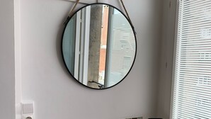 Een badkamer met een ronde spiegel, een witte wasbak, een zwarte kraan, een fles handzeep, een zwarte beker en een klein spiegeltje op een houten aanrecht.