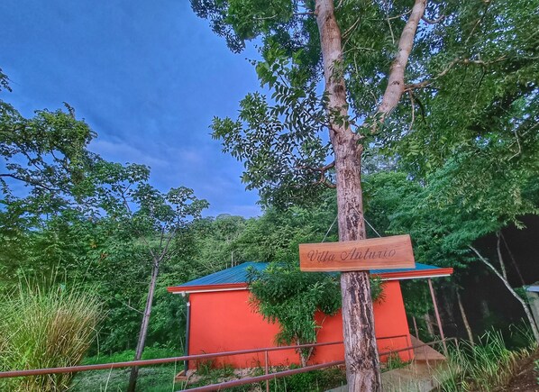 Een rood gebouw met een blauw dak, omgeven door weelderige begroeiing en bomen. Een houten bord met 'Villa Antwio' is zichtbaar.