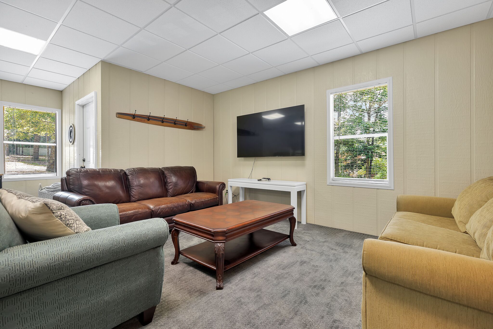 A living room with a brown leather couch, a green patterned sofa, a wooden coffee table, and a yellow armchair. A flat-screen TV is mounted on the wall, and there is a window with blinds.