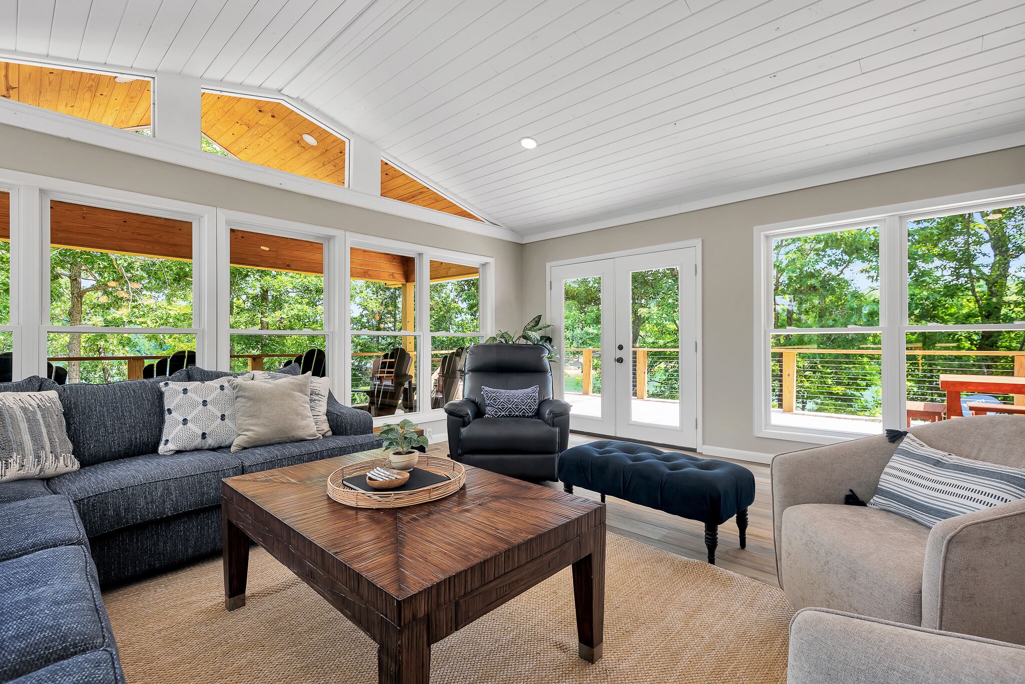 A spacious living room with a sectional sofa, a coffee table, and a black leather chair. Large windows provide views of the outdoors.