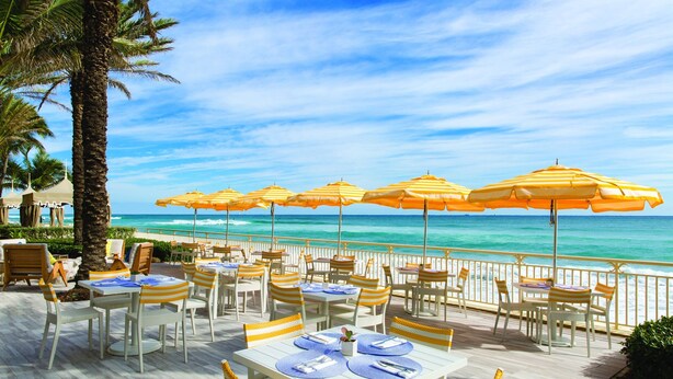 An outdoor dining area with tables and chairs, yellow striped umbrellas, and a view of the ocean. - Modern Romantic Tropical Poolside Retreat! (West Palm Beach)