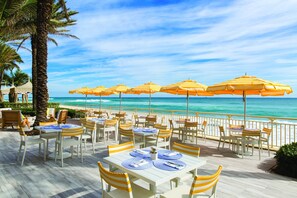 Ein Außenbereich zum Essen mit Tischen und Stühlen, gelb-weiß gestreifte Sonnenschirme und Blick aufs Meer.