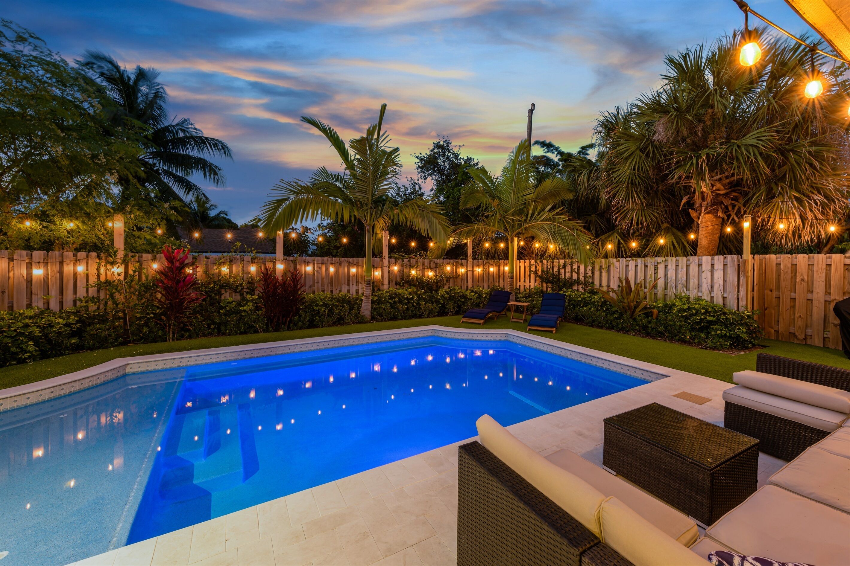 A swimming pool with a wooden fence, lounge chairs, and string lights.