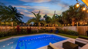 A swimming pool with a wooden fence, lounge chairs, and string lights. - Modern Romantic Tropical Poolside Retreat! (West Palm Beach)