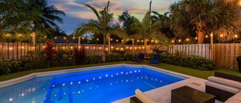 A swimming pool with a wooden fence, lounge chairs, and string lights.
