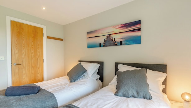 Une chambre d’hôtel avec deux lits, une porte en bois, un tableau représentant un coucher de soleil et une table de chevet avec une lampe.