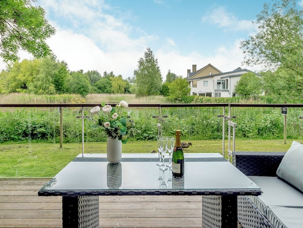 A glass table with a bottle of champagne, a vase of flowers, and wine glasses on a balcony overlooking a grassy area with trees and a house in the background.