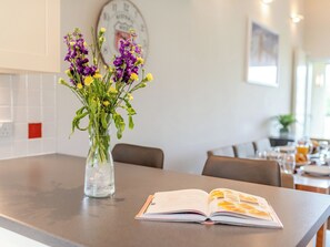 Una encimera de cocina con un florero transparente lleno de flores, un libro y un reloj en la pared.