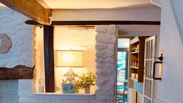 A cozy living room with a stone wall, a wooden beam, a sofa with cushions, and a small table with a lamp and a radio.