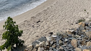 Ein Strand mit felsigem Vordergrund, sandigem Ufer und einer Hügelkulisse mit Vegetation und einem Gebäude.