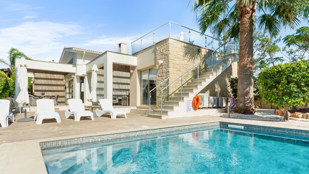 Ein modernes Haus mit Swimmingpool, weißen Liegestühlen und einer Treppe, die zu einem Balkon führt.