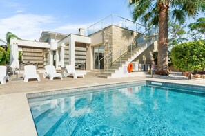 Une maison moderne dotée d’une piscine, de chaises longues blanches et d’un escalier menant à un balcon.